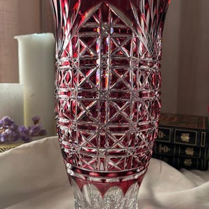 Puede incluir: Un jarrón de cristal cilíndrico y alto que presenta un color rojo rubí intenso sobre vidrio transparente. El exterior está tallado con un intrincado patrón geométrico de diamantes, estrías verticales en el borde y una base en estrella.
