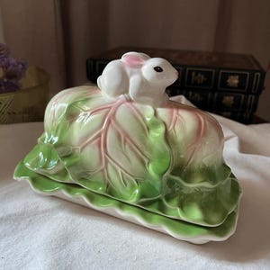 Peut inclure: Un beurrier en céramique représentant un lapin blanc sculpté aux oreilles teintées de rose, posé sur un couvercle en forme de chou. Les feuilles de chou sont peintes dans des tons vert clair et rose, avec une base aux bords festonnés.