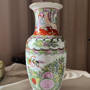 Peut inclure: Un vase en porcelaine blanche doté d'un bord évasé avec une finition dorée. Le col présente des personnages peints à la main dans un pavillon, tandis que le corps est orné de motifs floraux colorés, de feuillage vert et d'un petit oiseau.