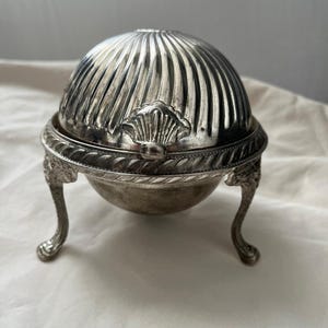 Pode incluir: Uma manteigueira de metal prateado com tampa abobadada e canelada, apresentando uma pega em forma de concha. A base é suportada por três pernas curvas e ornamentadas, e o rebordo é decorado com um padrão de corda torcida.