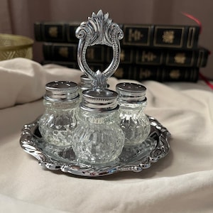 Puede incluir: Un juego de tres saleros y pimenteros de cristal transparente con tapas de metal plateado, dispuestos sobre una bandeja redonda de metal plateado a juego. La bandeja cuenta con un asa central decorativa con un diseño ornamentado.