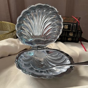 May include: A silver-toned metal clam shell dish with a hinged lid that opens to reveal a smooth, reflective interior. A small, matching silver-toned metal spoon with a shell-shaped bowl rests inside the open dish, which features a scalloped edge.