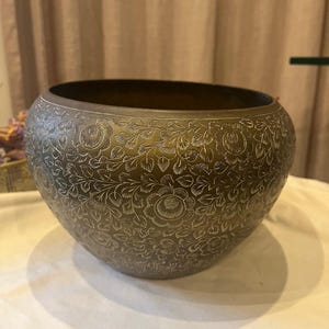 Pode incluir: Uma taça redonda de latão escuro com um intrincado padrão floral gravado que cobre toda a superfície exterior. O design consiste em flores detalhadas e videiras com folhas incisas no metal, criando um motivo botânico repetitivo.