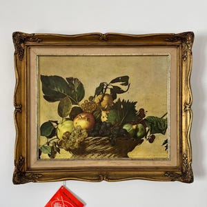 Pode incluir: Uma pintura a óleo numa moldura dourada decorativa representando um cesto entrançado cheio de vários frutos, incluindo uvas, maçãs e figos, acompanhados por folhas verdes. Uma etiqueta vermelha com texto branco e um logótipo está presa.