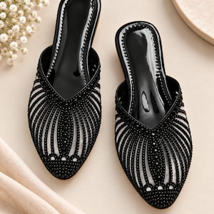 May include: A pair of black pointed-toe mules featuring a glossy black insole and a translucent upper adorned with intricate, radiating patterns of small black beads. The design includes a V-shaped vamp and a structured, slip-on silhouette.