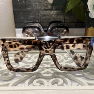 Peut inclure: Lunettes de vue carrées surdimensionnées avec un motif léopard marron et noir sur les montures épaisses en plastique. Les lunettes ont des verres transparents et un pont prononcé, présentant une forme angulaire audacieuse avec une finition brillante.