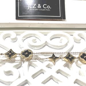 May include: A gold-tone chain bracelet featuring alternating black and gold-tone floral and circular charms. The jewelry is accompanied by a black tag with white text that reads JLZ & Co. JEWELLERY COLLECTION.