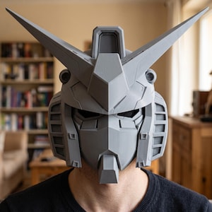 May include: A gray, 3D-printed robotic helmet featuring a sharp, angular design with two prominent, pointed antennae extending from the top. The helmet includes a central crest, recessed eye sections, and detailed ventilation grilles on the sides.