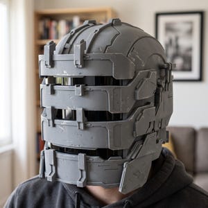 Pode incluir: Um capacete cinzento de cobertura total com um design segmentado de várias camadas, ranhuras horizontais para a viseira e um acabamento industrial desgastado. A estrutura inclui barras de suporte verticais na frente e detalhes.