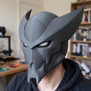 Puede incluir: Un casco integral de color gris oscuro con textura, que presenta prominentes aletas laterales curvadas hacia arriba y una placa frontal angular estilizada con aberturas oculares estrechas bordeadas en blanco. El casco tiene una estructura rígida.