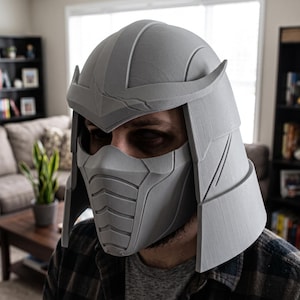Könnte beinhalten: Ein matter grauer 3D-gedruckter Helm mit segmentiertem Design, spitzem Stirnansatz, geschwungenen Seitenteilen und einer geformten Gesichtsmaske mit horizontalen Rillen über Nase und Mund, gefertigt mit deutlich sichtbaren Schichtlinien.