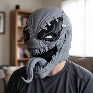 May include: A gray full-head mask featuring a textured surface, a wide mouth with sharp pointed teeth, and a long protruding tongue. The mask includes black eye patches with white accents and silver-colored rectangular stitches across the forehead.