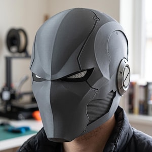 May include: A matte dark gray full-face helmet featuring a sleek, angular design with recessed eye sockets, white mesh eye coverings, and circular side ear pieces. The helmet has a textured surface with visible panel lines and a perforated chin section.