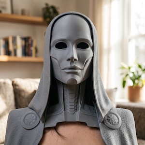 Op de afbeelding: Een grijs, 3D-geprint masker met een glad, mensachtig gezicht met holle oogkassen, een gestructureerd nekstuk en een textuurrijke kap. De kap is bij de schouders bevestigd met twee ronde medaillons, elk voorzien van een stervormig embleem.
