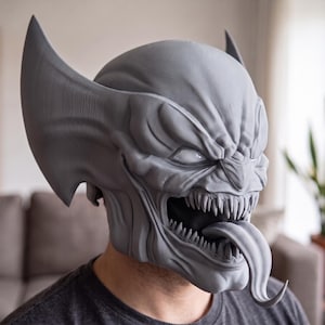 Op de afbeelding: Een grijs, 3D-geprint masker van een wezen met scherpe, puntige oren, een brede mond vol gekartelde tanden en een lange, uitstekende tong. Het masker heeft een getextureerd oppervlak en bedekt het bovenste deel van het hoofd en het gezicht.