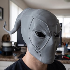 May include: A grey, full-head mask featuring a smooth, sculpted surface with defined facial contours, almond-shaped black eye lenses, and a prominent, curved, horn-like protrusion extending from the back. The mask includes stitched-like details.