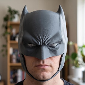 Puede incluir: Una máscara estilo Batman de color gris con orejas puntiagudas, una superficie texturizada y cubiertas de malla blanca para los ojos. La máscara cubre la frente y las sienes, dejando expuestos la nariz, la boca y la barbilla, con cejas esculpidas.