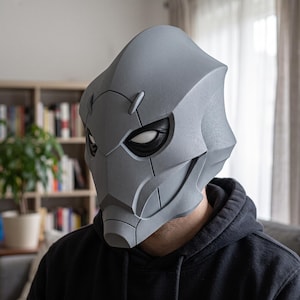 Può includere: Un casco integrale grigio dal design elegante e angolare, caratterizzato da una cresta centrale prominente, orbite nere incassate con lenti bianche e linee di pannello definite sulla superficie, che conferiscono un aspetto futuristico.