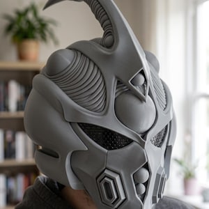 May include: A grey, full-head helmet featuring a prominent, curved horn-like protrusion at the top, intricate layered ridges, and a black honeycomb-patterned visor. The design includes geometric vents and circular accents across the front surface.
