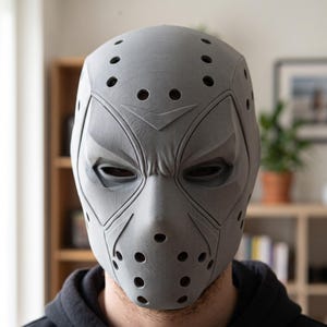 May include: A matte gray, full-face mask featuring a textured, 3D-printed finish. The design includes angular eye cutouts, a V-shaped detail on the forehead, and multiple circular ventilation holes across the brow, cheeks, and chin area.