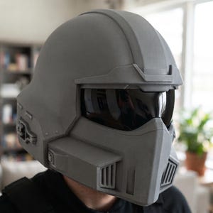 May include: A matte grey full-face helmet featuring a dark, reflective visor, a prominent brow ridge, and angular side vents. The helmet has a structured, geometric design with a protruding chin guard and integrated side panels.