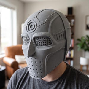 May include: A matte gray full-head mask featuring a mechanical design with a circular emblem on the forehead, a pointed nose piece, and mesh-covered eye openings. The lower section includes a perforated pattern, and the surface has molded rivets.
