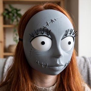 May include: A gray, full-face mask featuring large, round white eyes with black pupils, prominent black eyelashes, and raised stitch detailing across the forehead and cheeks. The mask has a smooth, matte finish and is secured with a black elastic strap.