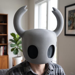 Pode incluir: Um capacete cinzento de superfície lisa com a forma icónica da cabeça da personagem Hollow Knight. O capacete tem duas grandes aberturas oculares ovais pretas e dois chifres curvos proeminentes que se estendem para cima a partir do topo.