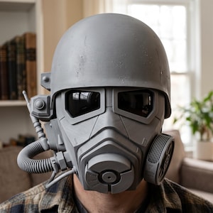 Puede incluir: Un casco táctico gris mate con diseño de cara completa, lentes oscuras, un puente nasal central prominente y un filtro circular en el lado izquierdo. Una manguera flexible conecta el filtro con la sección de la mandíbula inferior.