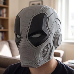 Puede incluir: Un casco de cabeza completa con un acabado gris claro texturizado y secciones oculares negras mate lisas. El diseño incluye detalles mecánicos intrincados a lo largo de la mandíbula y piezas circulares en las orejas, con un patrón de malla.