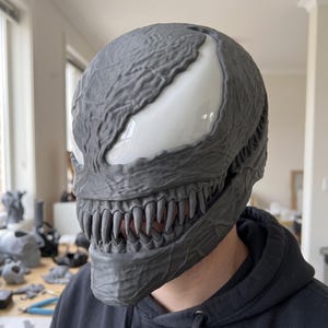 May include: A gray, textured full-head mask featuring large, smooth, white, almond-shaped eyes and a wide, menacing mouth filled with sharp, pointed teeth. The surface of the mask has a detailed, vein-like, organic pattern across the entire exterior.