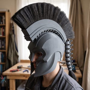 Peut inclure: Un casque de style spartiate gris mat doté d'une crête en brosse noire proéminente s'étendant de l'avant vers l'arrière. Le casque comprend une structure rigide segmentée en forme d'épine dorsale à l'arrière, ornée de poils noirs.