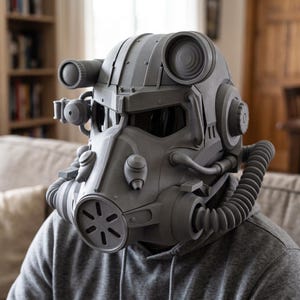 Pode incluir: Um capacete cinza fosco que cobre toda a cabeça, apresentando um filtro respiratório frontal proeminente, aberturas circulares nas laterais e uma viseira escura. O capacete inclui vários detalhes mecânicos, tubos e painéis estruturais.