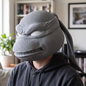 May include: A grey, textured, full-head mask resembling a turtle character with a prominent brow, a wide grin showing teeth, and a long, flowing head tie extending from the back. The mask features a pebbled skin texture and smooth, white eye slits.