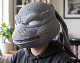 May include: A gray, textured, full-head mask resembling a turtle character with a prominent brow, a wide grin showing teeth, and a long, flowing head tie extending from the back. The mask features a pebbled skin texture and smooth, white eye slits.