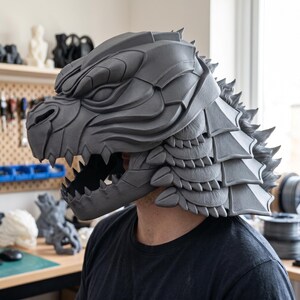 May include: A matte grey, full-head helmet sculpted in the likeness of a reptilian creature with sharp, jagged teeth, a prominent snout, and layered, scale-like plates covering the back and sides, creating a textured, armoured appearance.