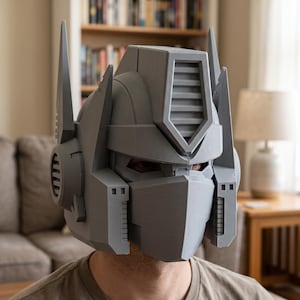 Op de afbeelding: Een grijze, 3D-geprinte robothelm met een geometrisch ontwerp, hoge, puntige zijantennes, een centrale geventileerde voorhoofdkam en een solide gezichtsplaat die de neus en mond bedekt, met zichtbare horizontale laagstructuren.