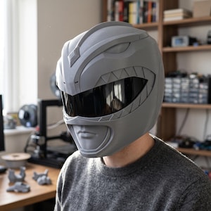 Könnte beinhalten: Ein mattgrauer Integralhelm mit einem stilisierten, von Dinosauriern inspirierten Design, einer markanten Mittelrippe, geformten Kieferpartien und einem breiten, dunkel getönten Visier, das den Augenbereich für ein einheitliches Aussehen abdeckt.