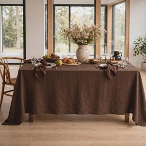 May include: A dark brown rectangular tablecloth draped over a dining table. The table is set with white plates, folded brown napkins, clear glassware, a bowl of grapes, a wooden board with bread, and a central vase filled with dried flowers.