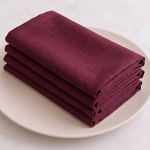 May include: A stack of four folded deep burgundy napkins made of a textured fabric, neatly arranged in a pile on top of a plain, round, light-coloured plate. The napkins feature a visible weave pattern and are stacked evenly to show their edges.