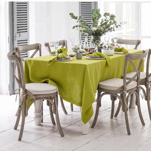May include: A rectangular lime green tablecloth draped over a dining table, accompanied by four wooden cross-back chairs with off-white seat cushions. The table is set with white plates, folded napkins, glassware, and a central floral arrangement.