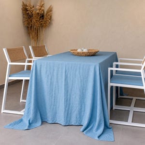 May include: A rectangular dining table covered with a solid light blue linen tablecloth, surrounded by three white metal chairs with light blue seat cushions and woven backrests. A round woven basket containing three white spheres sits on the table.