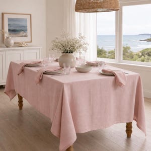 May include: A rectangular light pink tablecloth draped over a wooden table. The table is set with white plates, folded pink napkins, clear ribbed glasses, a central ceramic vase filled with white baby's breath flowers, and a small bowl.