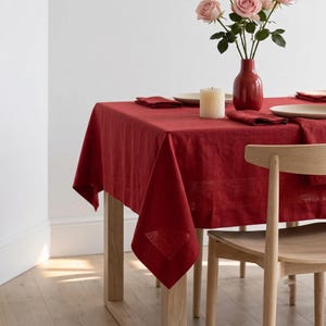 Puede incluir: Un mantel de lino rojo intenso sobre una mesa de madera, acompañado de un jarrón rojo con rosas rosadas, una vela blanca y servilletas rojas dobladas sobre platos blancos, junto a una silla de madera clara colocada en la mesa.