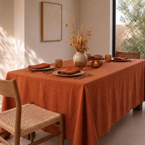 Op de afbeelding: Een rechthoekig roestoranje tafelkleed over een eettafel, gedekt met witte borden, gevouwen oranje servetten, amberkleurige glazen en goudkleurig bestek. Een crèmekleurige vaas gevuld met gedroogde gele bloemen staat in het midden.