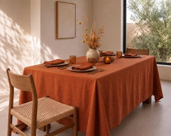 May include: A rectangular burnt orange tablecloth draped over a dining table, set with white plates, folded orange napkins, amber-coloured glassware, and gold-toned cutlery. A cream-coloured vase filled with dried yellow flowers sits in the centre.