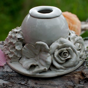 May include: A light gray ceramic candle holder featuring a central spherical vessel surrounded by sculpted floral elements including roses and hydrangea blossoms. The piece has a smooth, glossy finish and a circular base, crafted as a single unit.