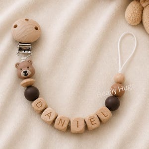 May include: A dummy clip featuring a round wooden clip, a brown bear-shaped bead, and wooden beads spelling the name DANIEL. The design includes dark brown, light brown, and natural wood beads, finished with a white cord loop.