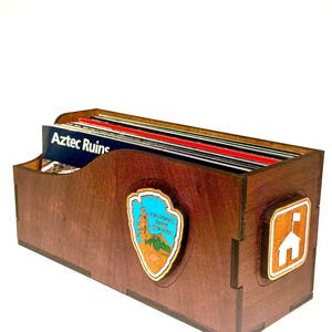 May include: A dark brown wooden storage box containing a collection of rectangular cards. The front of the box features a National Park Service arrowhead emblem and a side panel displays a white camping icon. One card is labeled Aztec Ruins.