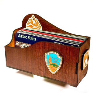 May include: A dark brown wooden organizer filled with National Park Service brochures, including one labeled Aztec Ruins. The front features a National Park Service arrowhead emblem, and the back panel displays a white mountain graphic sticker.
